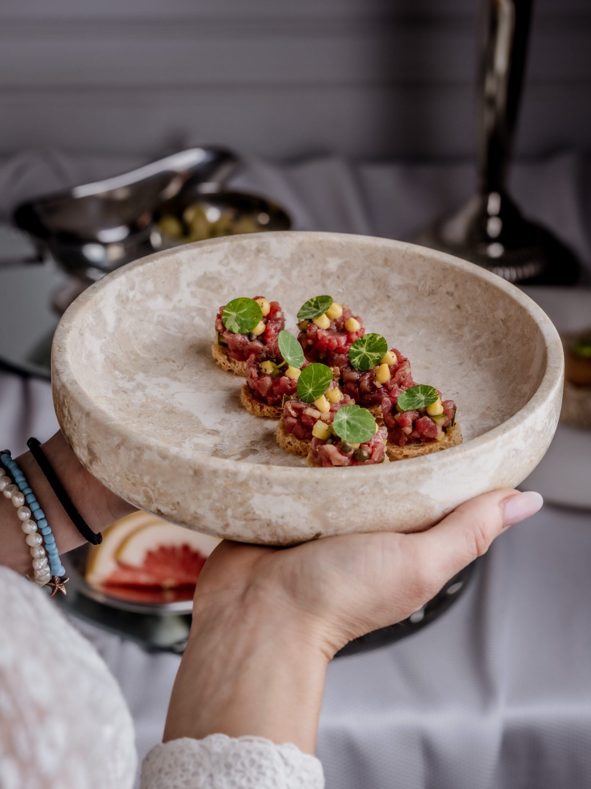 A close-up photograph of a hand holding a rustic, beige, stone-like bowl filled with five beef tartare canapés served on small toasts or crackers. The vibrant red tartare is garnished with yellow corn kernels and small green microgreens.